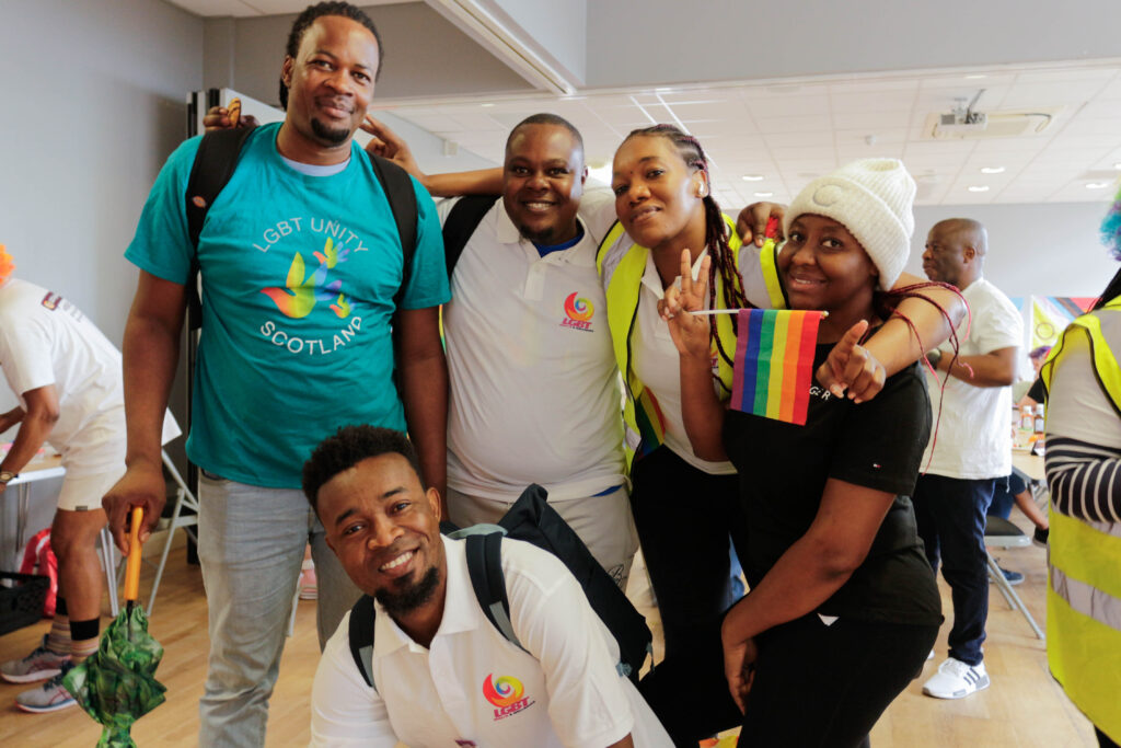 A diverse group of volunteers from LGBT Health and Wellbeing's Refugee Project smiling and posing together, proudly holding colorful rainbow flags in celebration.