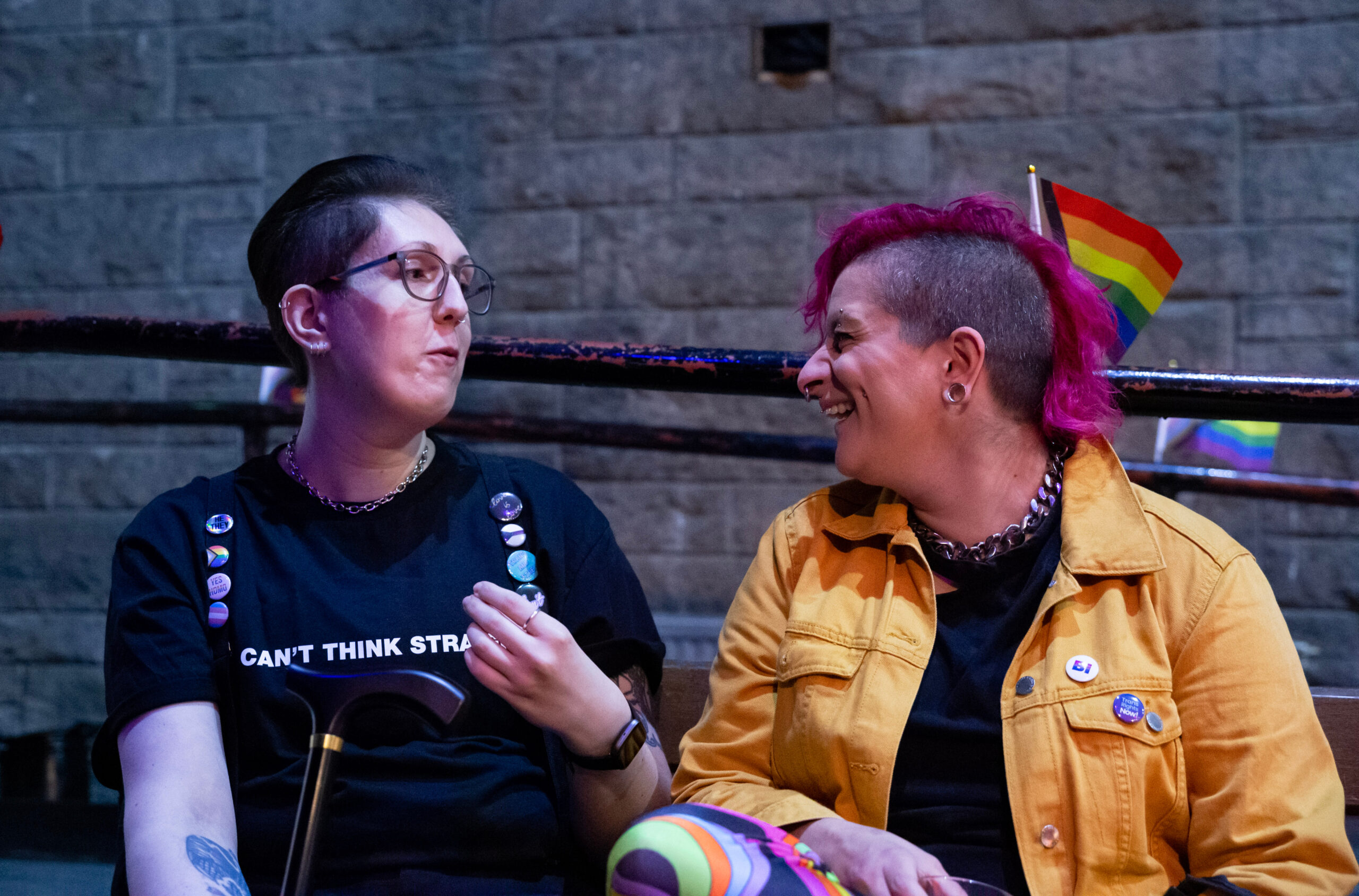 Two people sitting together, talking and smiling at an LGBT Health and Wellbeing event. One with short pink hair and a yellow jacket adorned with pins, the other wearing glasses and a black shirt that reads 'Can't Think Straight.'