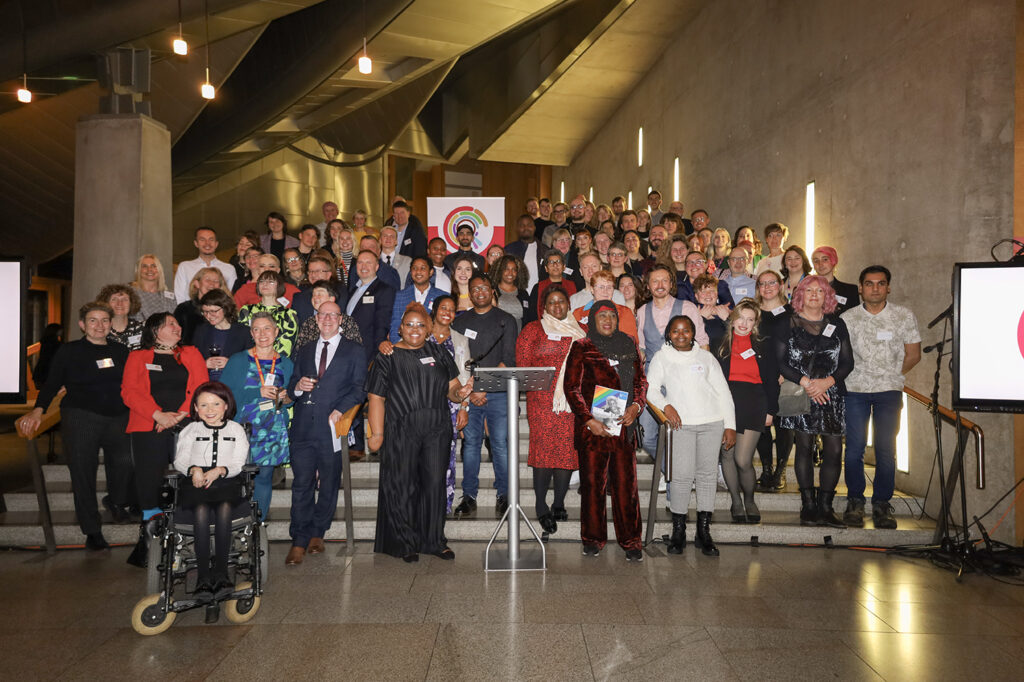A diverse group of people stands on stairs, smiling and posing next to LGBT Health and Wellbeing's banner.