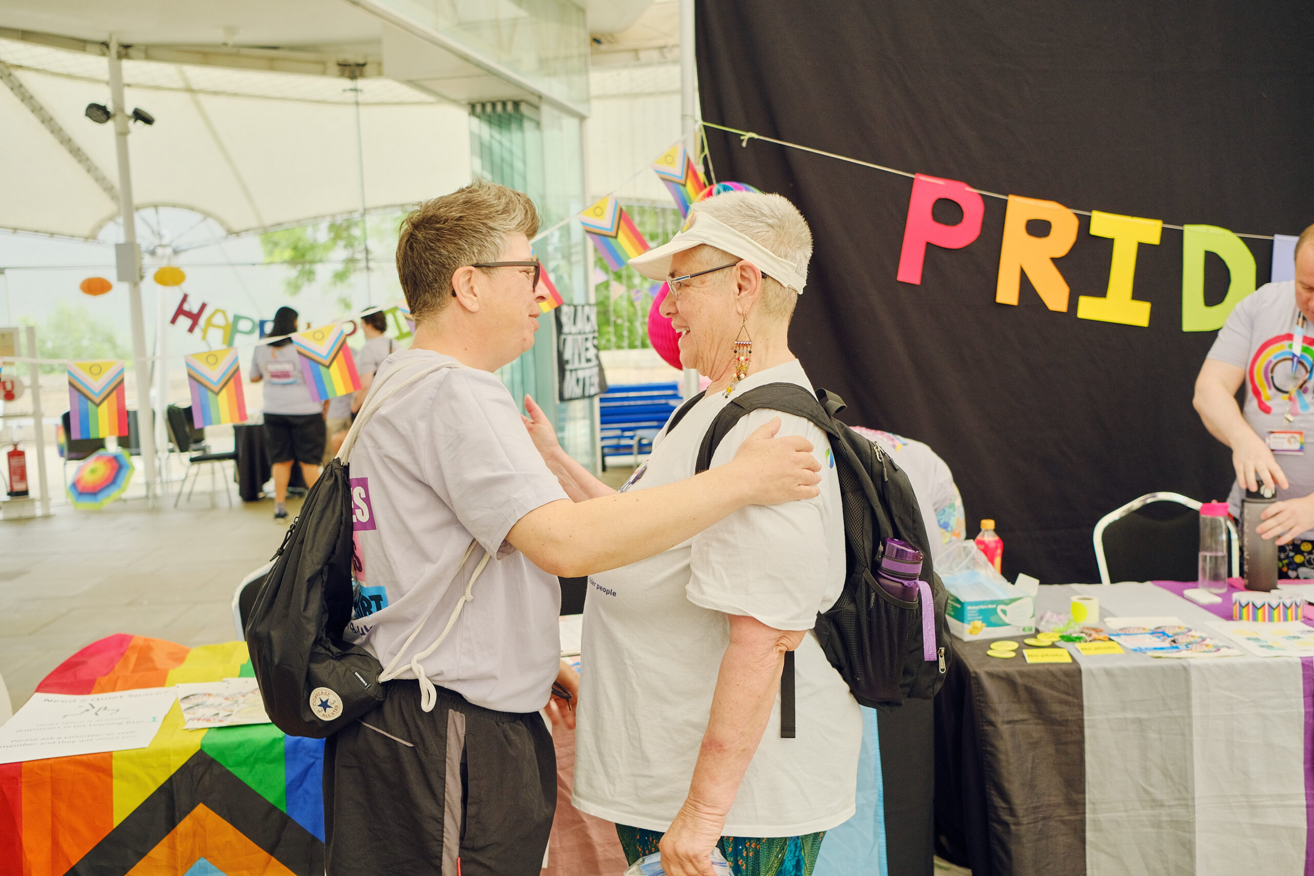 Two people stand close together at a Pride stall holding each other’s arms in a supportive gesture.