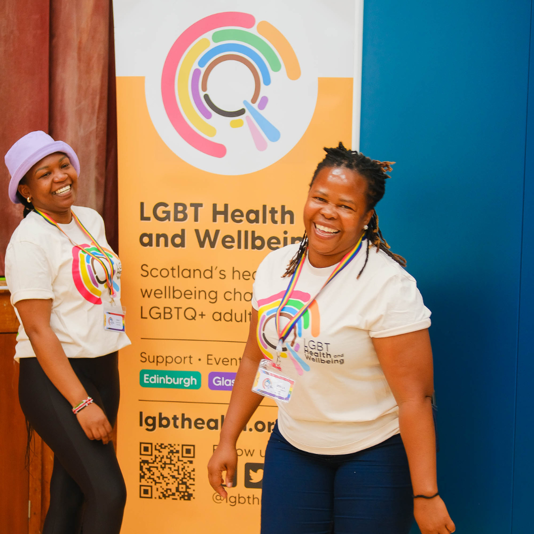 Two LGBT Health and Wellbeing team members standing in front of our logo banner